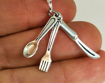 Może przedstawiać: Srebrzysty metalowy charms przedstawiający trzy miniaturowe sztućce: łyżkę, widelec i nóż. Elementy są przymocowane do małego okrągłego kółeczka połączonego z zapięciem typu karabińczyk, tworząc spójny dodatek o tematyce kuchennej.