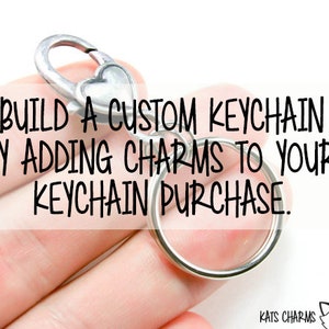 May include: A silver keychain with a heart-shaped charm and a round ring. The text "BUILD A CUSTOM KEYCHAIN BY ADDING CHARMS TO YOUR KEYCHAIN PURCHASE." is displayed on the image.