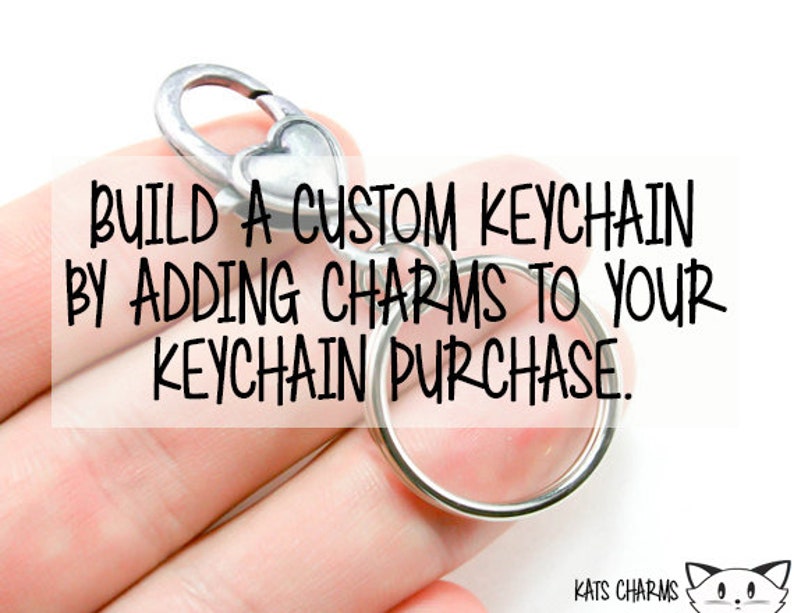May include: A silver keychain with a heart-shaped charm and a round ring. The text "BUILD A CUSTOM KEYCHAIN BY ADDING CHARMS TO YOUR KEYCHAIN PURCHASE." is displayed on the image.