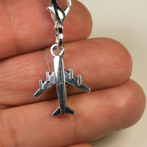 Könnte beinhalten: Ein silberfarbener Flugzeug-Charm mit einem Karabinerverschluss. Das Flugzeug hat detaillierte Flügel, einen Rumpf und ein Heck. Der Charm ist zum Anbringen an einem Armband oder einer Halskette gedacht. Der Charm wird in einer Hand gehalten.
