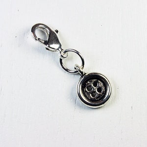 May include: A silver-toned charm featuring a button design. The charm has a small, round button with four holes in the center, attached to a clasp and a small ring. The clasp is open, ready to be attached to a necklace or bracelet.