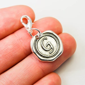 Op de afbeelding: Een zilverkleurige bedel met een karabijnsluiting. De ronde bedel lijkt op een zegel en heeft de letter "G" in het midden. De bedel heeft een getextureerde rand.