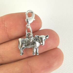 May include: A silver-colored cow charm with a lobster clasp. The charm is a detailed miniature of a cow, with spots and a textured surface. The charm is designed to be attached to a bracelet or necklace.