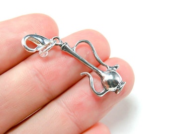 May include: Silver charm in the shape of a traditional Middle Eastern teapot with a decorative handle. The charm has a lobster claw clasp.
