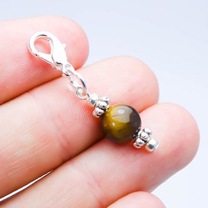 Op de afbeelding: Een zilverkleurige bedel met een karabijnsluiting en een ronde, gepolijste tijgeroogkraal. De kraal heeft een mix van bruine en gouden kleuren. Decoratieve zilveren accenten bevinden zich aan weerszijden van de kraal.