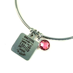 May include: Silver bangle bracelet with a square charm that reads "Grateful for where I'm at and excited for where I'm going". The charm is attached to a pink crystal.