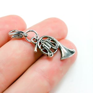 Könnte beinhalten: Silberner Anhänger in Form eines französischen Horns. Der Anhänger hat oben eine Schlaufe zum Anbringen an einem Armband oder einer Halskette.