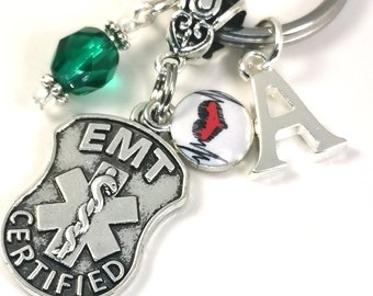 May include: Silver EMT keychain with a shield-shaped charm, a green faceted bead, and a letter "A" charm. The shield charm has the words "EMT CERTIFIED" and a medical symbol. A small charm features a red heart.