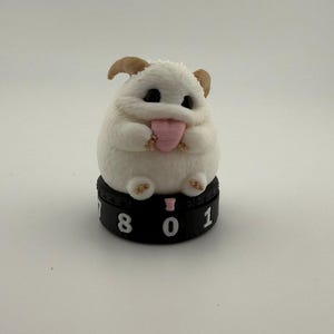 May include: A small white creature figurine with brown ears and large black eyes, holding a pink heart-shaped object. The figure sits atop a black circular base featuring white numbers including 8, 0, and 1, with a small pink icon above the zero.