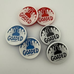 May include: A collection of six round, white buttons with the word "GOADED" in a bold, sans-serif font. The buttons feature a silhouette design in red, blue, and black. The buttons are arranged on a white surface.