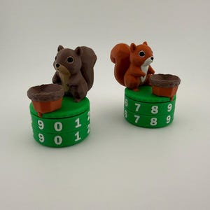 May include: Two squirrel-shaped counters, one brown and one orange, each standing on a green circular base with rotating numbered dials. The dials display white numbers ranging from zero to nine, allowing for manual tracking of scores or tallies.