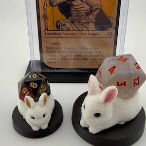 Könnte beinhalten: Eine Magic: The Gathering-Karte mit Cadira, Caller of the Small, einem Orc Ranger. Zwei weiße Hasenfiguren mit Würfeln obenauf befinden sich vor der Karte. Die Würfel sind rot und schwarz.