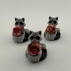 Könnte beinhalten: Drei graue Waschbärfiguren, die jeweils einen cupcake-förmigen Würfelhalter mit einem rot-weiß marmorierten Würfel halten. Die Würfel zeigen die Zahl 13 und andere Zahlen in Gold. Die Figuren stehen auf schwarzen kreisförmigen Sockeln.