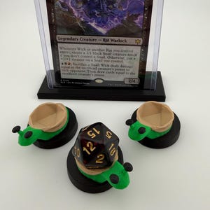 May include: A Magic: The Gathering card, "Wick, the Whorled Mind," in a protective case, is displayed above three green and black dice holders shaped like turtles. A large, multi-sided die rests in one holder.