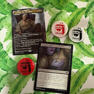 May include: An assortment of Magic: The Gathering cards and pinback buttons. The cards feature vampire-themed artwork and text, including "Astarion, the Decadent" and "Vampire's Kiss." The buttons display a carton of blood graphic with the word "Blood."