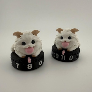Peut inclure: Deux figurines blanches et duveteuses avec des oreilles fauves et des langues roses, chacune posée sur une base circulaire noire. Les bases présentent des chiffres blancs et un petit indicateur rose, servant de compteurs de points de vie.