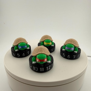 May include: Four Teenage Mutant Ninja Turtle figurines. Each turtle has a different colored mask and is atop a black base with white numbers. The turtles have tan shells and green bodies. The bases are round.
