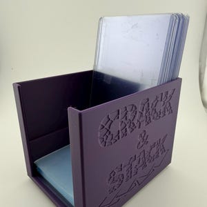 Può includere: Una scatola di stoccaggio viola con le parole "Crack & Stack" in rilievo sul lato. La scatola è aperta, rivelando pile di custodie per carte di plastica trasparente e custodie per carte blu all'interno. La scatola è realizzata in materiale solido.
