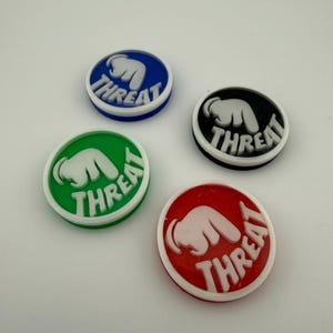 May include: Four circular shoe charms in blue, green, black, and red. Each charm features a white raised graphic of a hand pointing downward with the word THREAT embossed in white capital letters across the bottom of the circular surface.