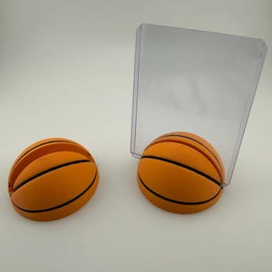 May include: Two orange basketball-shaped card holders with black stripes. One holder is holding a clear plastic card. The holders are designed to resemble basketballs, with the classic orange color and black lines.