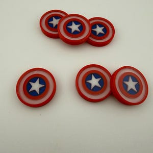 以下が含まれることがあります： キャプテン・アメリカの盾型の装飾品がいくつかあります。各盾は赤い縁、白い内側の円、青い円、中央に白い星が特徴です。白い表面に配置されています。
