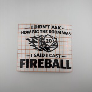 May include: A square sticker with a grid pattern and black text that reads "I DIDN'T ASK HOW BIG THE ROOM WAS. I SAID I CAST FIREBALL." A black and white die with the number 20 is in the center, with flames.