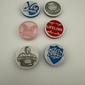 May include: Six round, colorful tokens with various designs and text. The tokens include "HEX PROOF", "VIGILANCE", "FLYING", "LIFELINK", "INDESTRUCTIBLE", and "SHIELD". Each token has a unique color scheme and graphic.