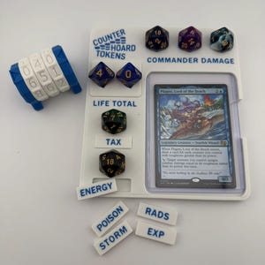 May include: A white plastic card game organizer with dice and tokens. The organizer has slots for dice, a card, and small tokens labeled "ENERGY", "POISON", "STORM", "RADS", and "EXP". A blue and white counter is on the left.