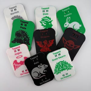 May include: A selection of gaming tokens in white, green, and black, featuring illustrations of creatures like a rabbit and a dragon. Each token is labelled with the words "Tapped" and "Un-Tapped". The tokens are designed for a tabletop game.