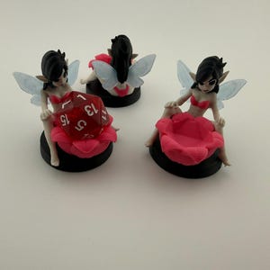 May include: Three figurines of fairies with black hair, blue wings, and red tops. Each fairy sits on a pink flower atop a black base. One holds a red, multi-sided die. The other two are empty.