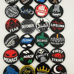 May include: A collection of twenty-four circular black tokens featuring various white, red, blue, and yellow graphic icons and text labels including Goaded, Vigilance, Shield, Double Strike, Shroud, Ward, Hexproof, Trample, Fear, Defender, Intimidate, Lifelink, Deathtouch, Flying, Decayed, Indestructible, Menace, Reach, Finality, First Strike, Skulk, Ring Bearer, Monarch, and Night.