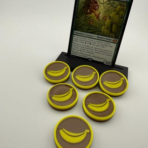 May include: A Magic: The Gathering card, "Kibo, Uktabi Prince," is displayed in a black stand, accompanied by six brown and yellow banana-themed tokens. The card features a monkey illustration and game text.