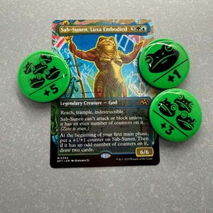 May include: A Magic: The Gathering card, "Sab-Sunen, Luxa Embodied," with four green frog-themed counters. The card features a frog-like deity. The counters have values of +1, +3, and +5. The card text describes the creature's abilities.