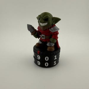May include: A green goblin figurine with a brown beard, wearing a red tunic and silver armor, holding a small gray dagger. The figure stands atop a black cylindrical life counter dial featuring white numbers 9, 0, and 1 printed on the side.