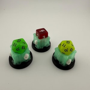 May include: Three miniature tabletop game pieces. Each piece features a colored die atop a translucent green cube, mounted on a black circular base. One die is red, and two are green and yellow.
