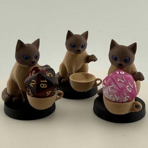 Peut inclure: Trois figurines de chat aux yeux bleus et au pelage brun, chacune tenant une tasse à thé. Une tasse à thé contient un dé bordeaux et or, une autre une tasse à thé simple, et la troisième un dé rose et blanc. Toutes sont sur des socles noirs.