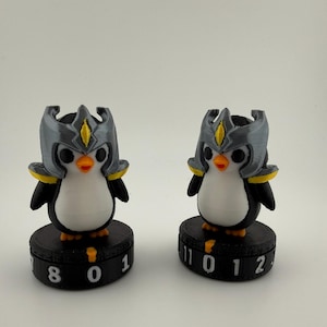 May include: Two penguin figurines wearing grey helmets with yellow accents, standing on black circular bases. Each base features a rotating dial with white numbers, including 8, 0, 1, 11, and 2, designed to track game scores or statistics.