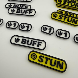 May include: An assortment of black and white and yellow gaming tokens. The tokens have the words "BUFF" and "STUN" in black, with a white border. Some tokens have a "1" with a symbol.