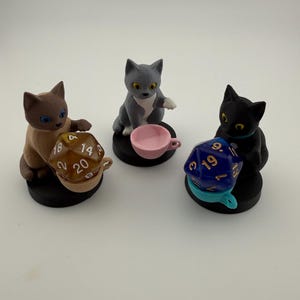 Puede incluir: Tres pequeñas figuras de gatos sobre bases circulares negras. Un gato beige sostiene un dado de veinte caras marrón en una taza, un gato gris se sienta junto a una taza rosa y un gato negro sostiene un dado de veinte caras azul en una taza azul claro.