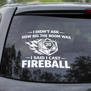 May include: A white vinyl decal on a car window. The decal features the text "I DIDN'T ASK HOW BIG THE ROOM WAS - I SAID I CAST FIREBALL" with a stylized D20 die graphic. The car is dark grey.