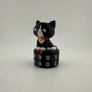 May include: A black and white cat figurine with a red collar and a small gold bell, sitting atop a black cylindrical mechanical counter. The counter features two rows of white numbers, including 8, 9, 0, and 1, printed on its rotating surface.
