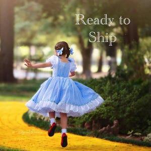 May include: A young person in a blue and white gingham dress with white lace trim and red shoes is walking on a yellow brick road. The dress has white sleeves and a square neckline. The text "Ready to Ship" is in the upper right corner.