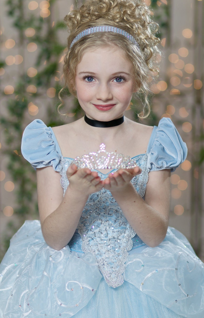 Pu&ograve; includere: Una giovane persona indossa un abito da principessa azzurro con una tiara d'argento. L'abito ha maniche a sbuffo e un corpetto con paillettes. La tiara &egrave; tenuta tra le mani ed &egrave; adornata da gemme scintillanti. Lo sfondo &egrave; sfocato con luci bokeh.