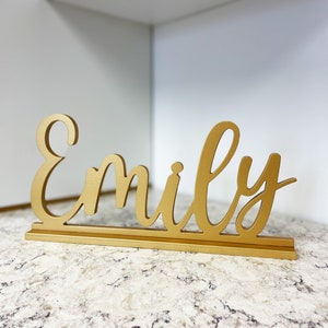 May include: Gold-colored wooden sign with the name "Emily" written in a cursive font.