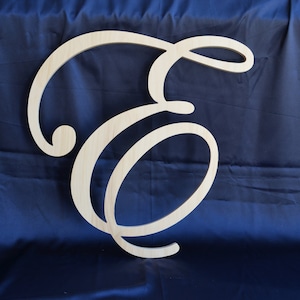 May include: A large, light-colored wooden cutout of the uppercase letter E, featuring a decorative, flowing cursive script font with smooth, rounded edges and a natural wood grain finish visible throughout the surface of the letter.
