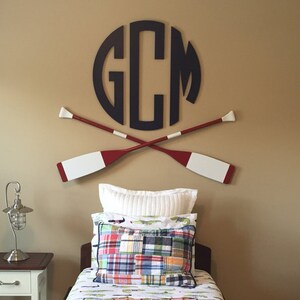 May include: A navy blue monogram with the letters "GCM" in a circle, with two red and white oars crossed behind it. The monogram is on a beige wall above a twin bed with a white and green plaid bedspread and a white pillow with a plaid pillow on top.