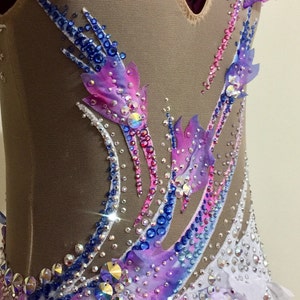 May include: A close-up of a rhythmic gymnastics leotard. The leotard features a nude mesh base with intricate embellishments. It is adorned with a gradient of pink, purple, and blue fabric accents, and sparkling rhinestones. The design includes feather-like patterns.