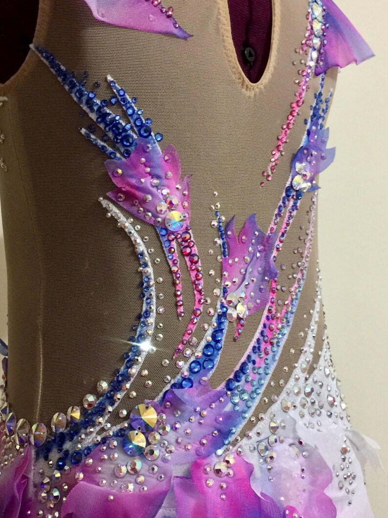 May include: A close-up of a rhythmic gymnastics leotard. The leotard features a nude mesh base with intricate embellishments. It is adorned with a gradient of pink, purple, and blue fabric accents, and sparkling rhinestones. The design includes feather-like patterns.
