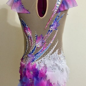 May include: A gymnastics leotard with a nude mesh base, adorned with a vibrant array of colors. The design features a gradient of pink, purple, and blue, accented with sparkling rhinestones and layered petal-like fabric.