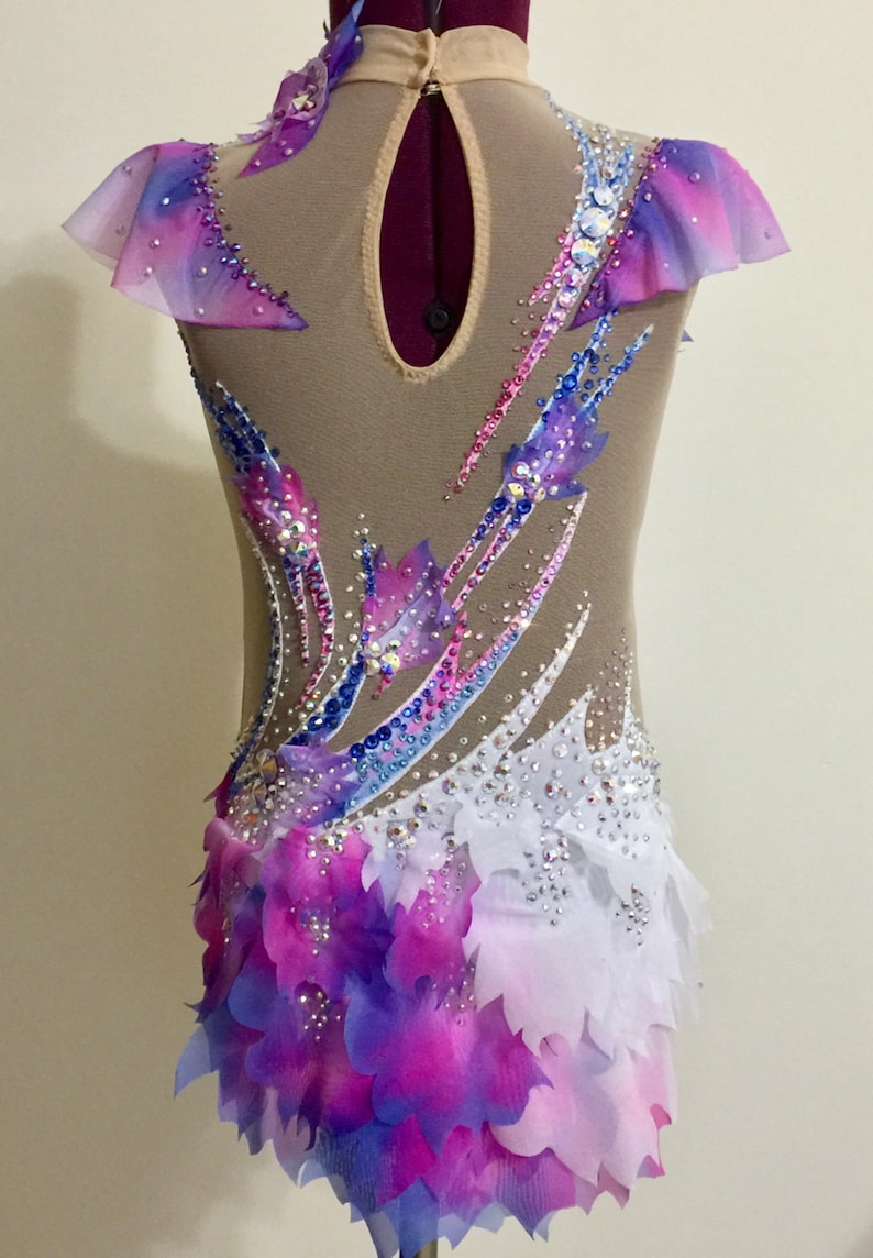 May include: A gymnastics leotard with a nude mesh base, adorned with a vibrant array of colors. The design features a gradient of pink, purple, and blue, accented with sparkling rhinestones and layered petal-like fabric.